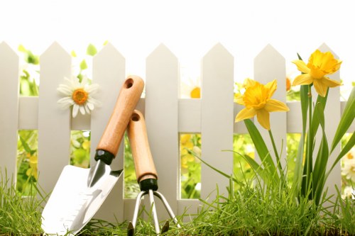 A close-up view of a well-maintained garden border featuring vibrant yellow daffodils and white daisies growing alongside a white picket fence, with a lush green lawn in the foreground. Gardening tools including a hand trowel and a pruner with wooden handles are placed on the grass, suggesting ongoing garden maintenance or planting activity. The scene is set outdoors on a bright, sunny day, with natural light illuminating the flowers and greenery. This image captures elements typical of a residential garden in Southwark, highlighting the tidy layout with flower beds adjacent to a grass lawn, and reflects the professional gardening services provided by Gardening Services Southwark aimed at enhancing outdoor spaces in London suburbs.