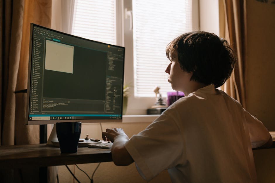 A woman with short dark hair sits at a wooden desk in a well-lit room, working on a computer with a large monitor displaying coding or software development. Behind her, natural light filters through p