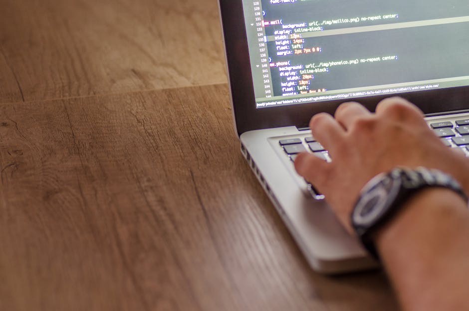 A close-up view of a person's hand with a wristwatch, typing on a silver laptop placed on a wooden surface. The laptop screen displays lines of coding or programming text with various lines of HTML or
